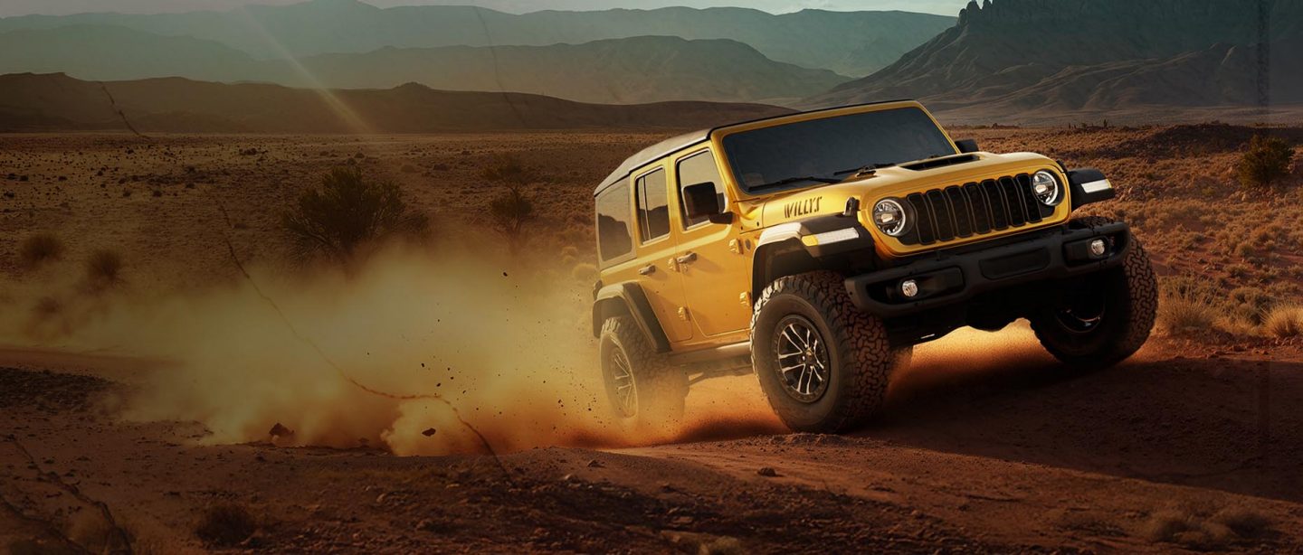 A passenger-side front angle of a gold 2026 Jeep Wrangler Willys 392 ascending a trail in the desert.