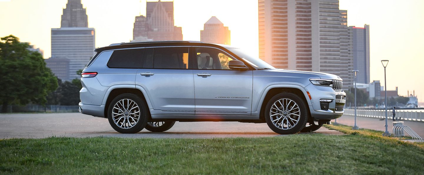 A profile view of a 2021 Jeep Grand Cherokee L Summit Reserve parked near a waterfront promenade with a cityscape at sunset in the background.